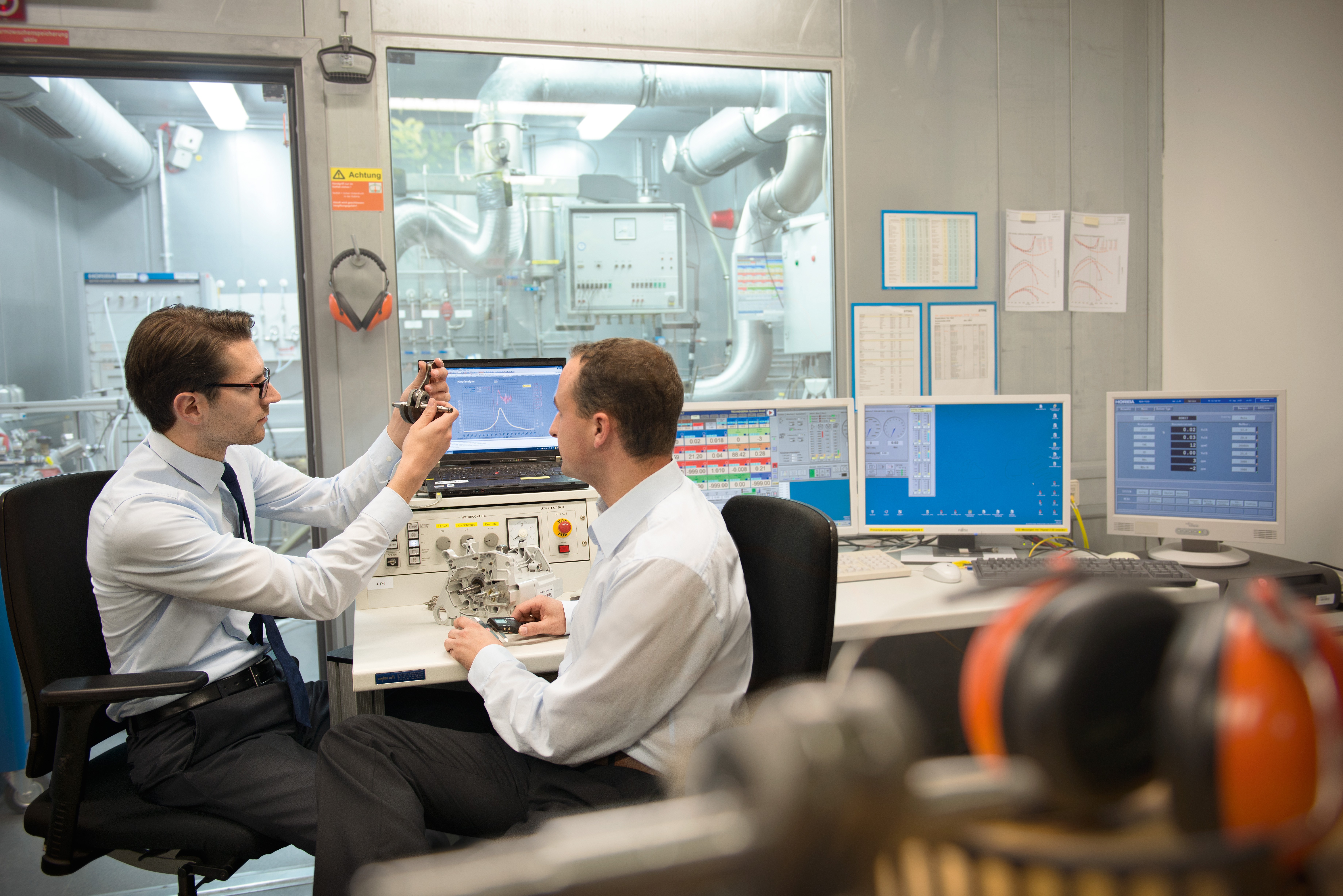 Two men in research and development sit in a laboratory at a desk with several monitors and examine part of a product