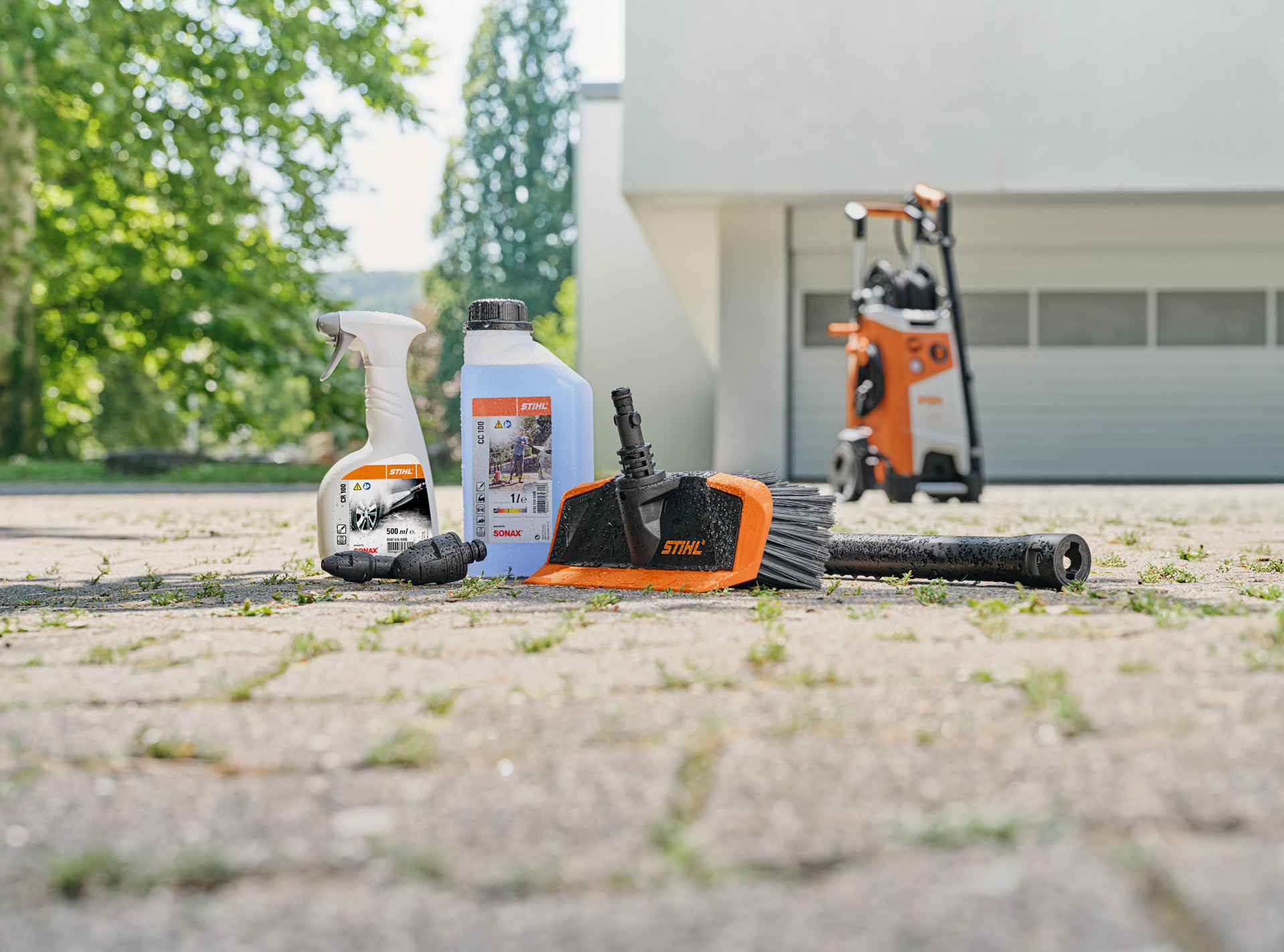  STIHL rim cleaner, vehicle shampoo and wash brush on the ground in front of a white garage. A STIHL high-pressure cleaner in the background